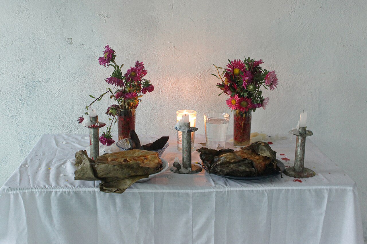 Altar sencillo de Hanal Pixán con mantel blanco, flores, velas y jícaras